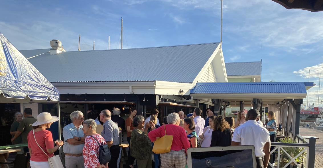 A daytime gathering on the marina square outside Aziana — guests on the cobblestones, the building behind, boats in the marina beyond