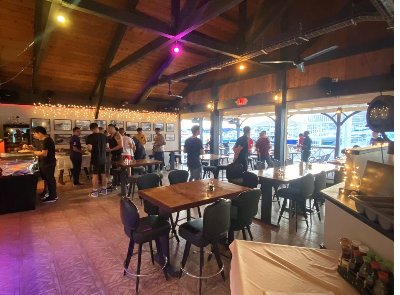 A private event indoors at Aziana — purple uplighting on the vaulted wood beams, string lights, the marina beyond the windows